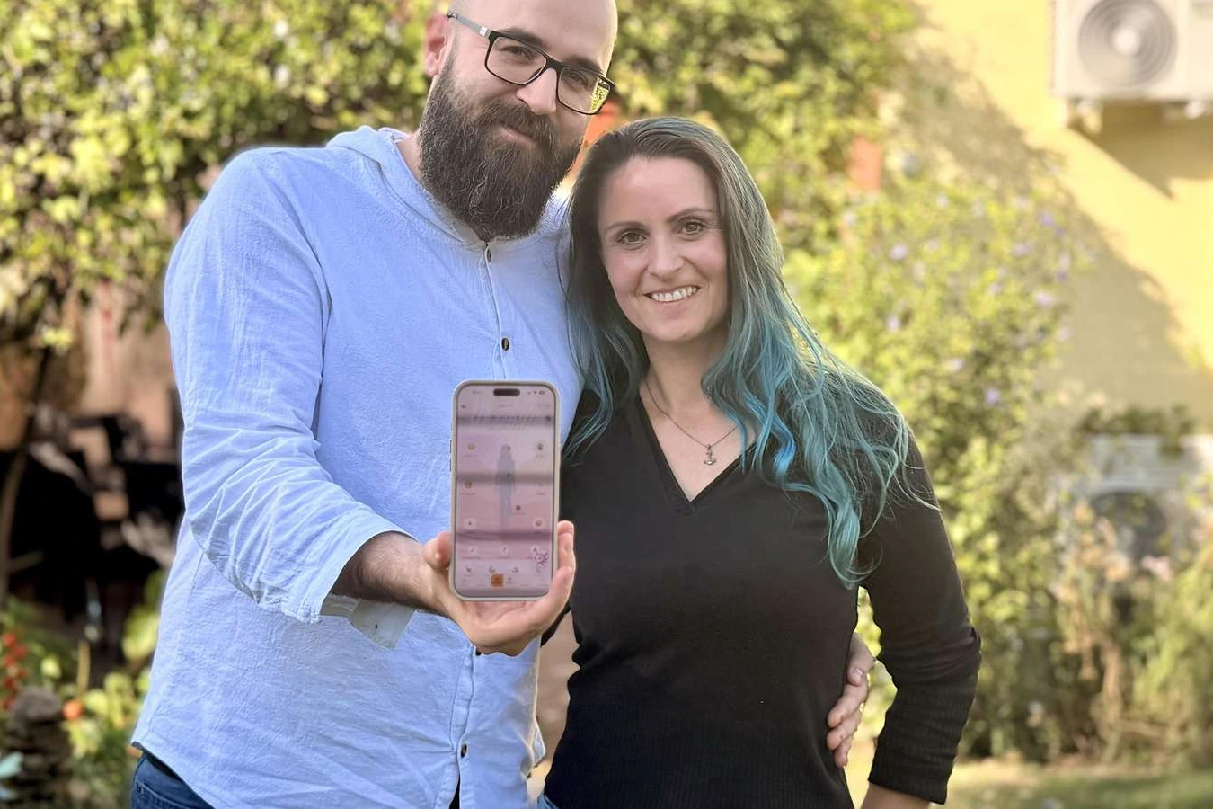 Cindy and Daniel März stand together. Daniel holds up a smartphone showing the BodySeasons app.